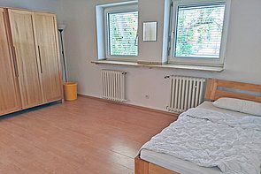 Zimmer mit 2 Fenstern, einem Bett und einem Kleiderschrank.