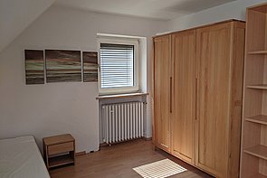 Zimmer des Hufeland-Hauses für Freiwillige, mit Fenster, Bett, Kleiderschrank, einem offenen Schrank und einem Nachttisch.
